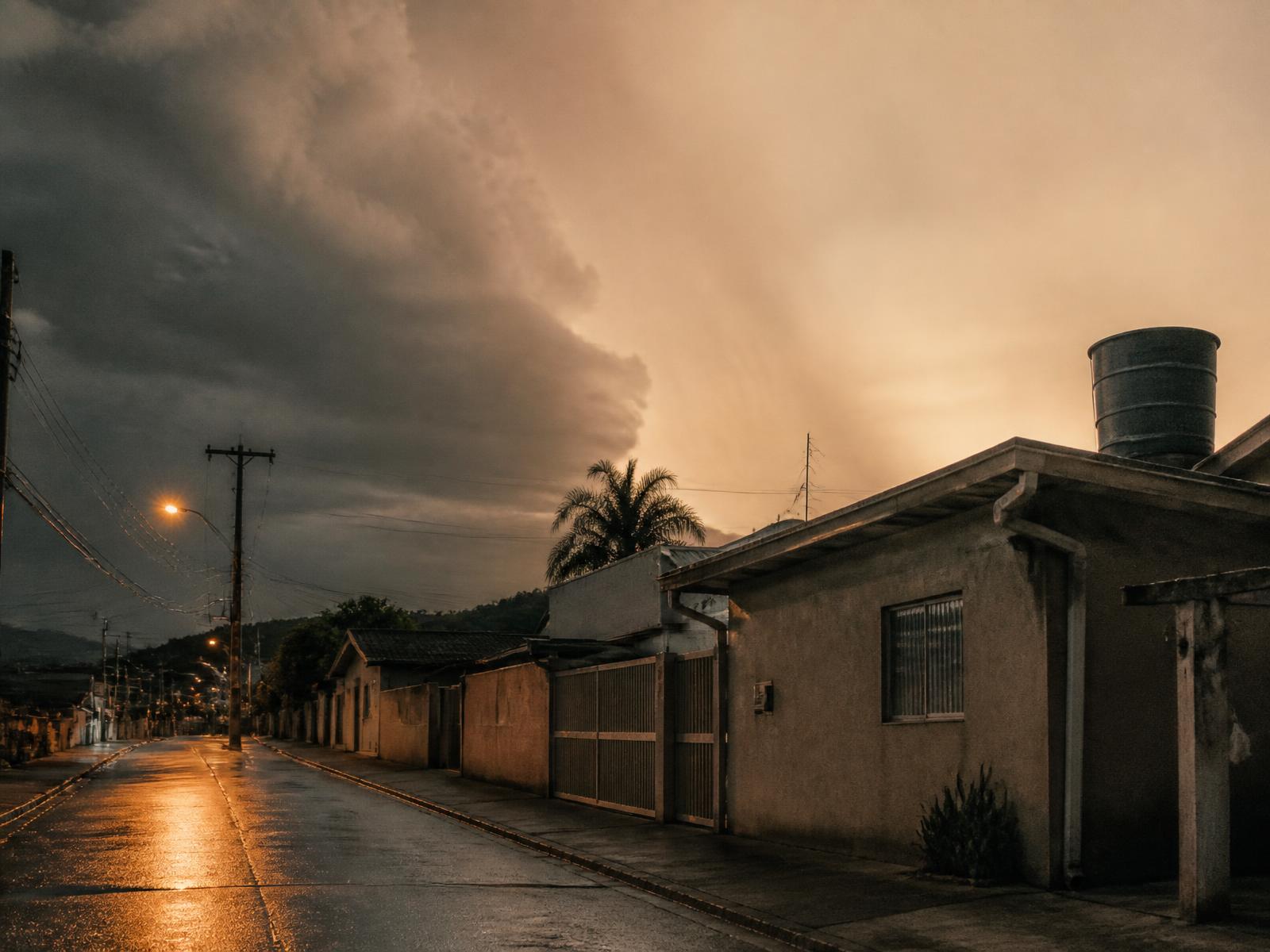 Rua residencial brasileira ao entardecer sob céu de tempestade, com caixa d'água e calhas visíveis